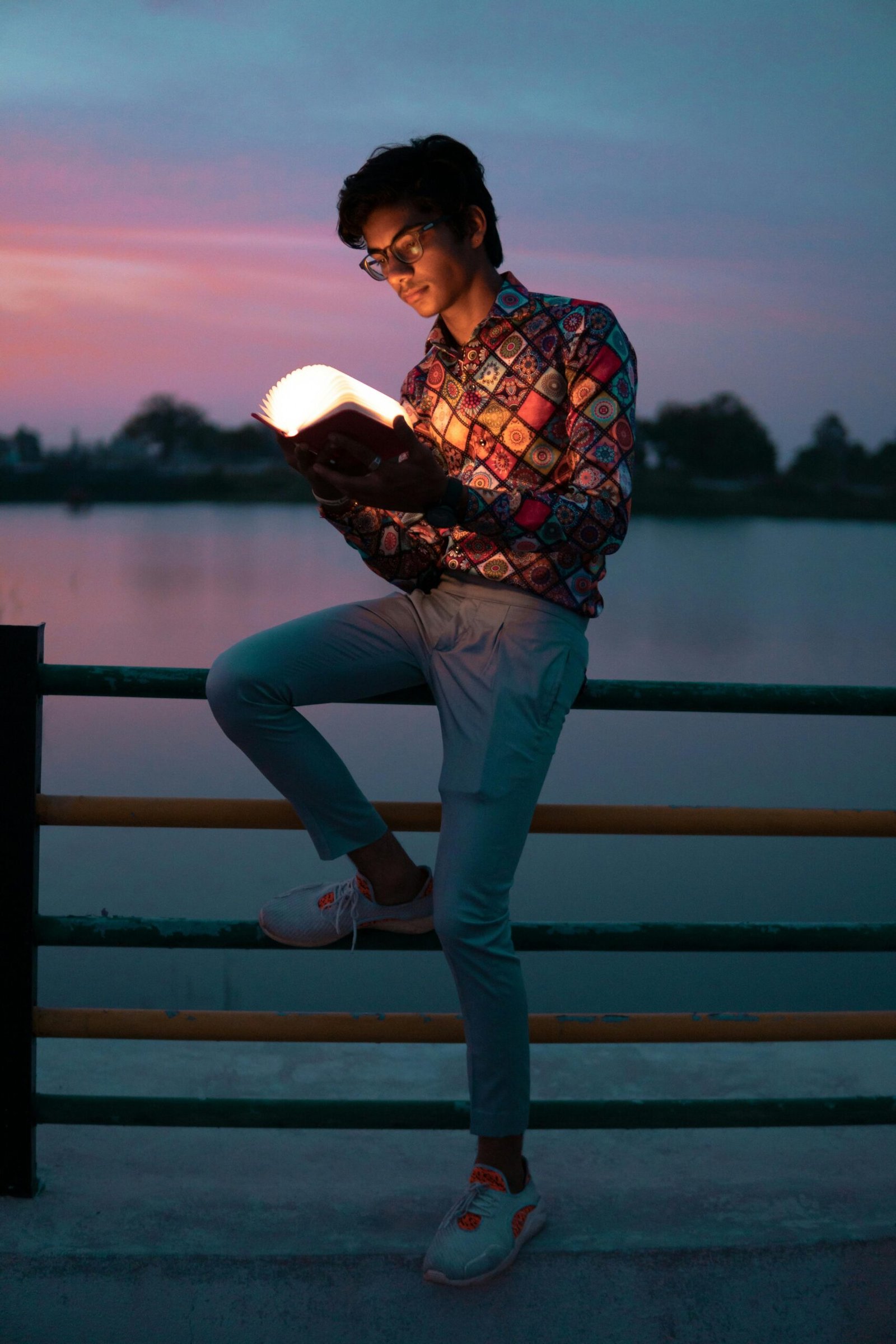 A young man reads by a lake at sunset, enjoying a moment of leisure and relaxation.